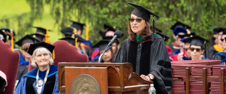 Commencement speaker speaking behind a podium and microphone with faculty and administrators watching