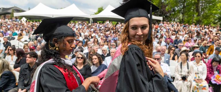 two graduates walking in front of audience. One graduate is fixing the others collar