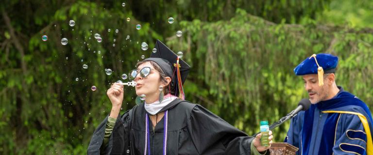 Commencement speaker speaking behind a podium and microphone behind a graduate blowing bubbles