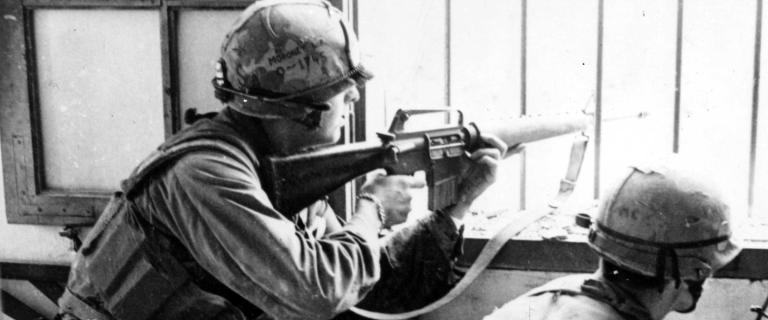 Black and white photo of two crouched soldiers looking out a window, one is point rifle out of the window.