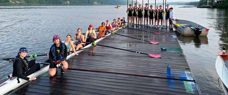 A dock on a lake with hills in the background. In the water a rowing scull is filled with people and five people on the dock holding a paddle.