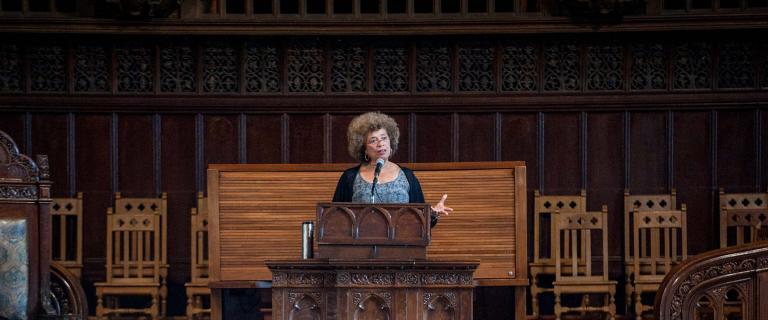 Angela Davis standing at a podium in front of a microphone wearing a gray and black shirt and black sweater. 