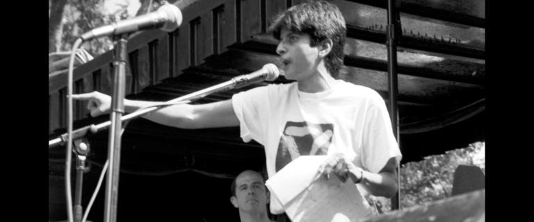 Black and white photo of Urvashi Vaid standing at a podium in front of 2 microphones pointing in front at Union Square Park in NYC.