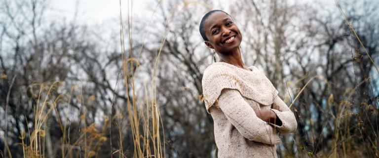 Amina Luqman-Dawson smiling with arms folded standing in front of a treeline.