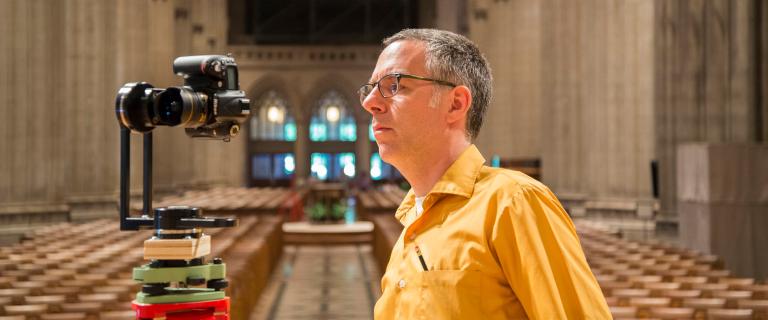 In a nave, a man with short dark hair, wearing glasses and a bright yellow short sleeve shirt, is looking intently at a camera view screen with pews in the background.