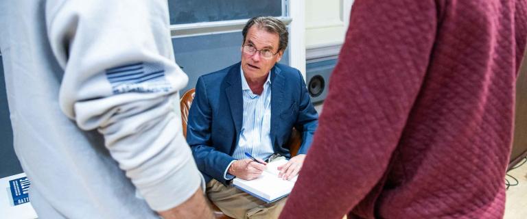 A person sitting in a chair signing a book and talking to two standing people.