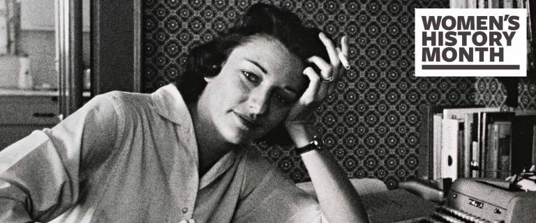 Rollie McKenna sitting with her left hand on her head, holding a cigarette, with books and a typewriter in the background, in black and white. 