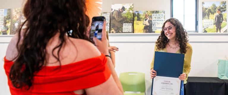A person holding a diploma award smiling while another person holds a phone taking their picture.