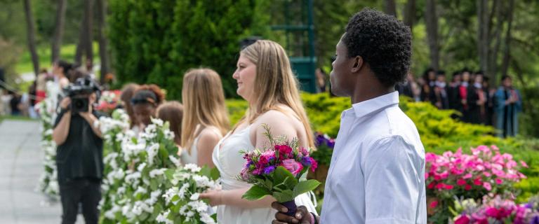 People in a line at a graduation ceremony holding flowers and looking on.