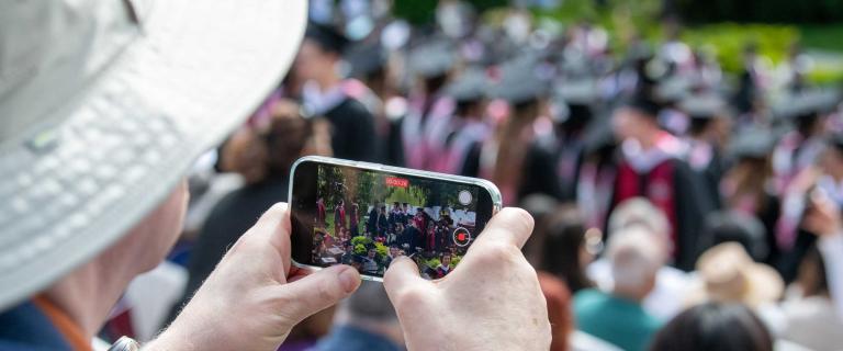 Person with a hat and a phone recording video of the stage at a graduation ceremony.