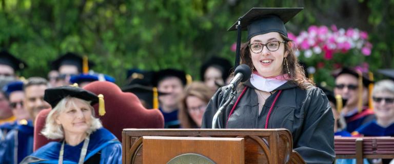 Person in graduation ceremony attire speaking on stage behind a microphone.