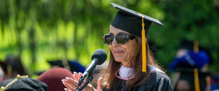 A graduation speaker in ceremonial attire behind a microphone clapping and smiling.