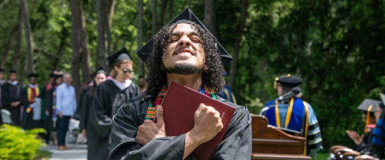 Person in graduation ceremony attire clutching their diploma and with eyes closed and a elated expression.