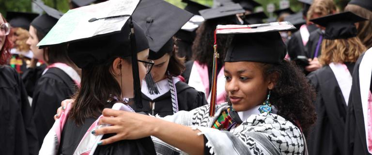 Two people in graduation ceremony attire. One is adjusting the other's collar.