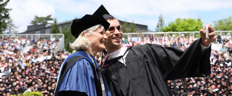 Two people in graduation ceremony attire on stage standing closely and posing for a selfie.