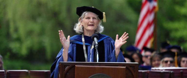 Person in graduation ceremony attire speaking on stage behind a microphone and a lectern.