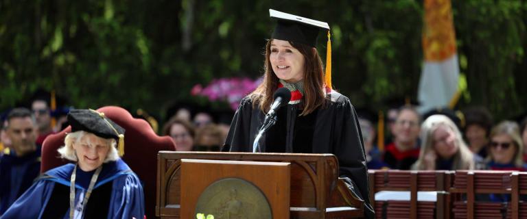 Person in graduation ceremony attire speaking on stage behind a microphone and a lecturn.