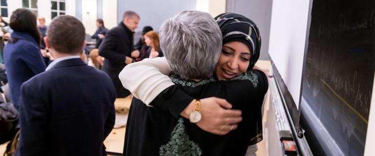 Two people hugging in a conference room with people and desks in the background.