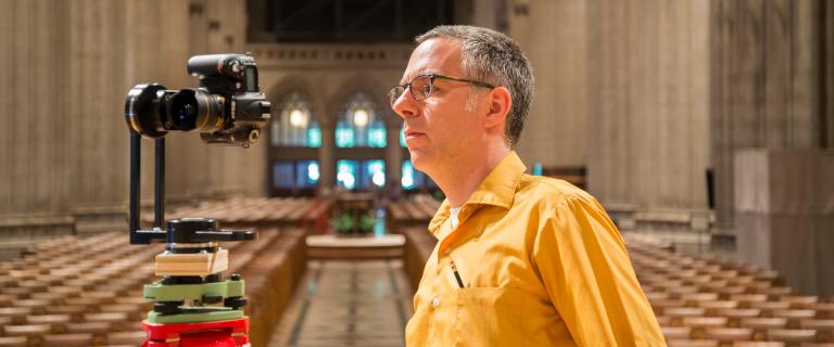 Andrew Tallon wearing a yellow shirt inside the Washington National Cathedral with a camera on a tripod. 