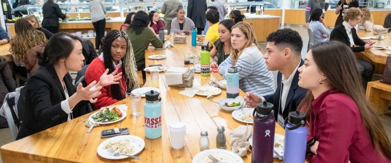 A group of people seated at a large table in a cafeteria talking over a meal.