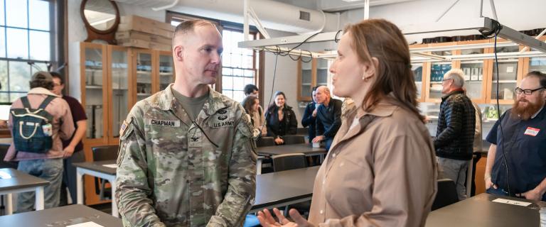 Two people standing in a classroom speaking to each other with other people and classroom tables in the background. One of the people speaking is wearing a camouflage, military uniform. 