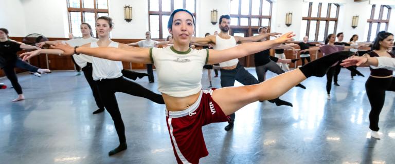 A dancer on point with their arms spread with other dancers doing the same in a studio.