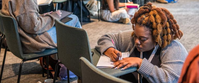 Person kneeling and writing in a notebook placed on a chair.