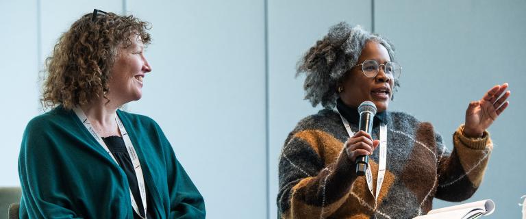 Two people sitting on a stage with one speaking into a microphone to an audience.