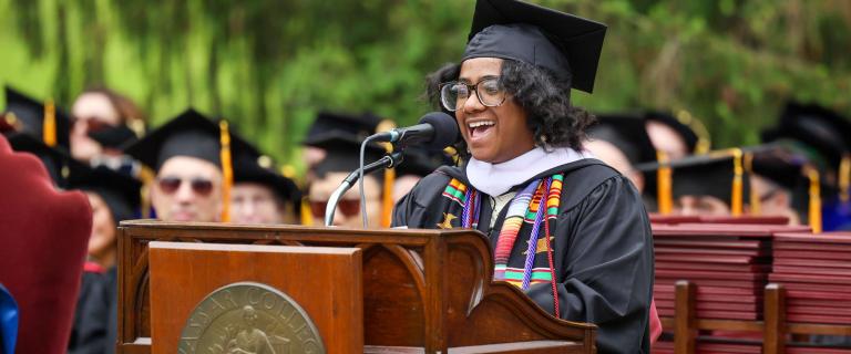 A speaker in graduation regalia stands at a wooden podium engraved with the Vassar College seal, addressing an outdoor audience through a microphone. The attire includes a black gown, mortarboard, white academic hood, and multicolored honor cords draped over the shoulders. Rows of seated individuals in black caps and gowns fill the background, with a stack of red diploma folders visible to the right. The setting is surrounded by green trees under soft daylight.