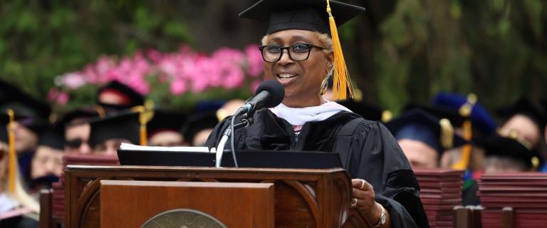 A speaker in academic robes and a mortarboard stands at a wooden podium bearing the Vassar College seal, addressing an audience with a microphone. The attire includes a black gown with a white academic hood, and a paper is held in one hand. Behind the speaker, seated individuals in black graduation caps fill the frame, with pink blossoms and trees in the background. A neatly stacked row of red diploma folders appears to the side.