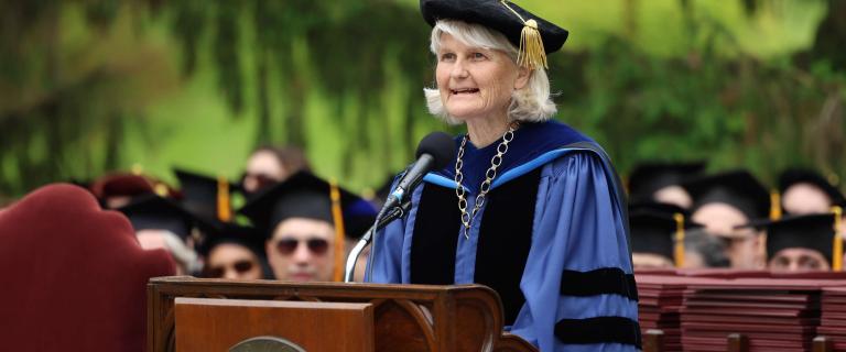 A figure dressed in a blue doctoral gown with black velvet panels and a gold tassel tam stands at a wooden podium marked with the Vassar College seal. A ceremonial chain hangs over the gown, and a microphone is positioned in front. In the background, individuals in black academic caps are seated outdoors, framed by trees and diffused daylight. A stack of red diploma folders is visible to the right of the podium.