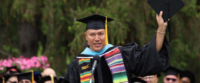 A speaker wearing a black graduation gown, colorful stole, and light blue hood raises a mortarboard in one hand while standing at a microphone. The podium bears the Vassar College seal, and seated individuals in caps and gowns are visible behind. The background includes green trees and clusters of pink flowers. A stack of red diploma covers sits on the stage nearby.