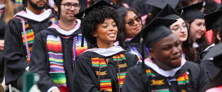 Graduates in black caps and gowns are seated closely together in rows during an outdoor ceremony. Several wear white academic hoods and brightly colored stoles in kente or striped serape patterns. One graduate near the center is looking slightly upward and smiling, while others nearby appear attentive or relaxed. The background is densely filled with more seated individuals in academic dress.