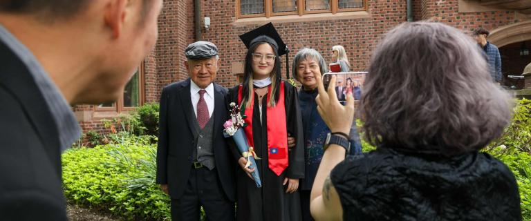 A person in graduation regalia stands between two others in front of a red brick building with leaded glass windows. The graduate wears a black gown, mortarboard, white academic hood, and a red stole featuring a small flag, while holding a rolled document wrapped in ribbon and a bouquet of flowers. A fourth person takes a photo of the group using a smartphone, with part of their head and hand visible in the foreground. Green shrubs and a stone walkway frame the lower part of the scene.