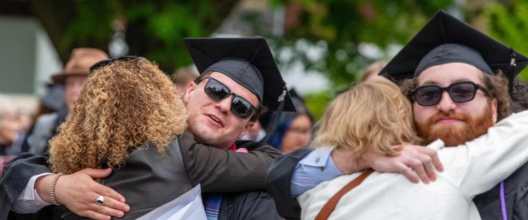 Two graduates wearing caps, gowns, and sunglasses embrace guests during an outdoor celebration. The group is surrounded by a crowd of people in a green, tree-lined setting. One graduate hugs someone with curly hair, while the other is mid-embrace with a person in a light-colored jacket. The mood is festive, with blurred faces and activity visible in the background.