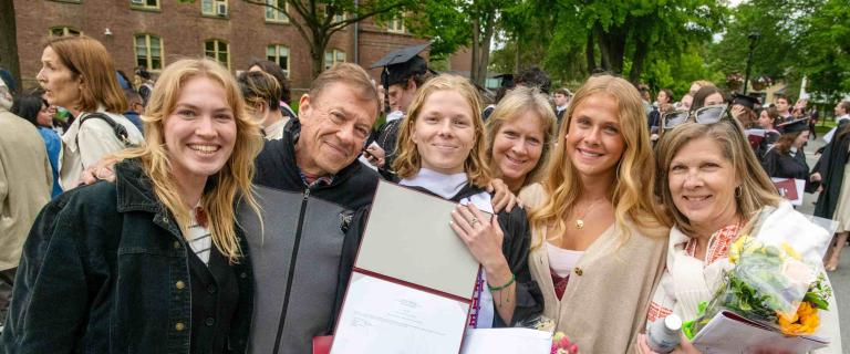 Six people pose closely together outdoors, with one holding an open diploma folder in graduation robes. The group stands smiling in front of a crowd, with red brick buildings, trees, and other graduates visible in the background. Some individuals hold flowers and printed programs. The lighting is bright and overcast.