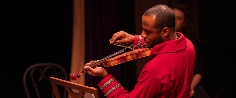 Person on a dark stage in a red jacket playing the violin.