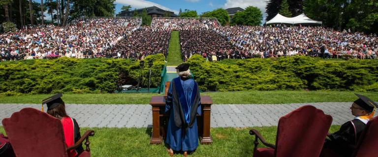 A commencement speaker in cap and gown speaking behind a lectern before a large outdoor audience.