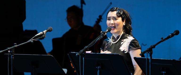 Performer sings into a microphone on stage with musicians in silhouette behind.