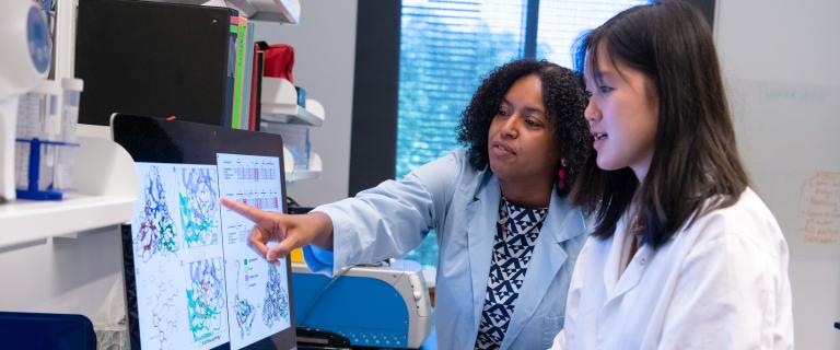 Two people wearing lab coats examine 3D protein structure models on a large computer monitor in a laboratory. One person points at the screen while the other operates the computer.