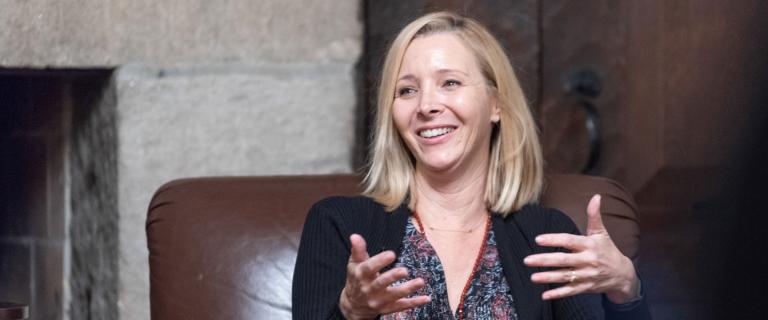 Photo of Lisa Kudrow seated and smiling in a chair speaking while using her hands.