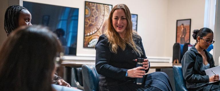 Dr. Michelle Monje ’98, a person with long blond hair and a dark dress, sits in a room talking to some other people.