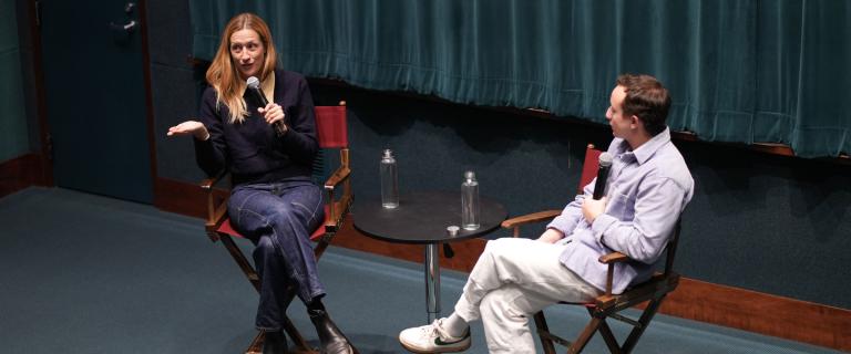 Two people seated in directors chairs on a stage holding microphones having a conversation.