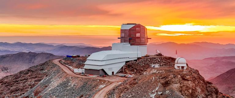 Vera C. Rubin Observatory sits atop a rocky mountain ridge at sunset, with vibrant orange and yellow hues lighting the sky.