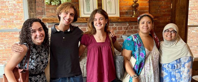 Five people standing side-by-side with their arms around each other in a classroom smiling.