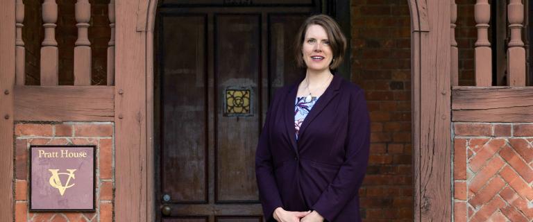Callista Isabelle in a purple blazer stands in front of a wooden doorway at Pratt House, with a sign displaying the Vassar College logo on the brick wall beside her.