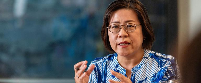 A person wearing glasses and a blue-and-white patterned blouse speaks while gesturing with their hands during a discussion.