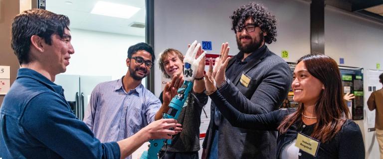 A group of onlookers checking out a robotic hand, held by a person, offerring a high five.