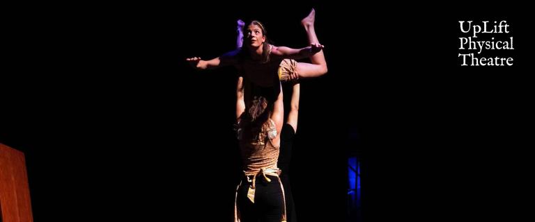 A dark stage with an actor holding a dancer above their head like they are flying.