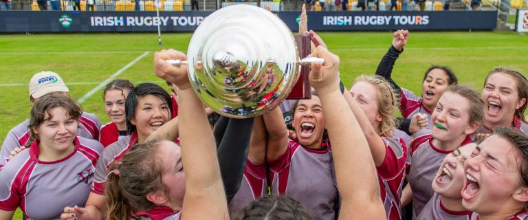 Vassar College Women’s rugby team in maroon and gray jerseys celebrating on the field, holding up a large silver trophy together with excitement.